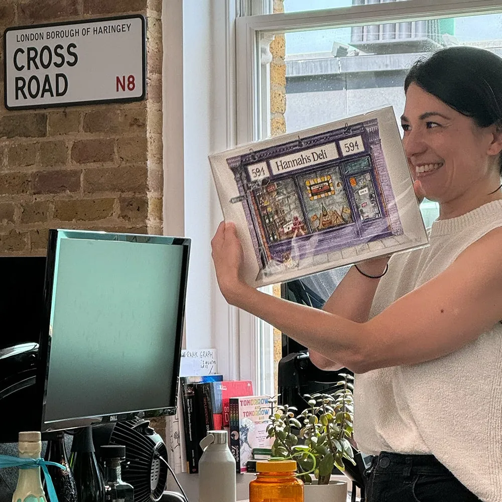 A delighted woman in an office holding up a framed illustration of Hannah's Deli shop front