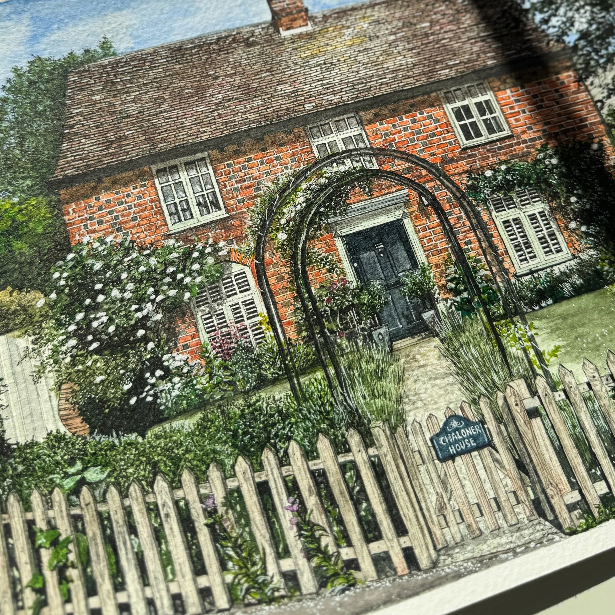 Ink and watercolour drawing of a red brick cottage with a rose arch, climbing plants and a white picket fence