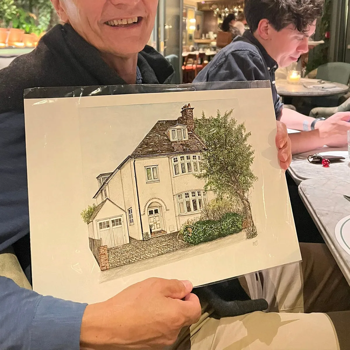 A smiling customer holding up his framed ink and watercolour portrait of a white house
