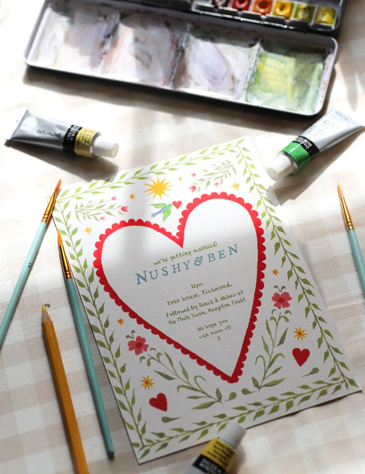 Illustrated wedding invitation with a red scalloped heart and a leafy green border, lying among paints and pencils