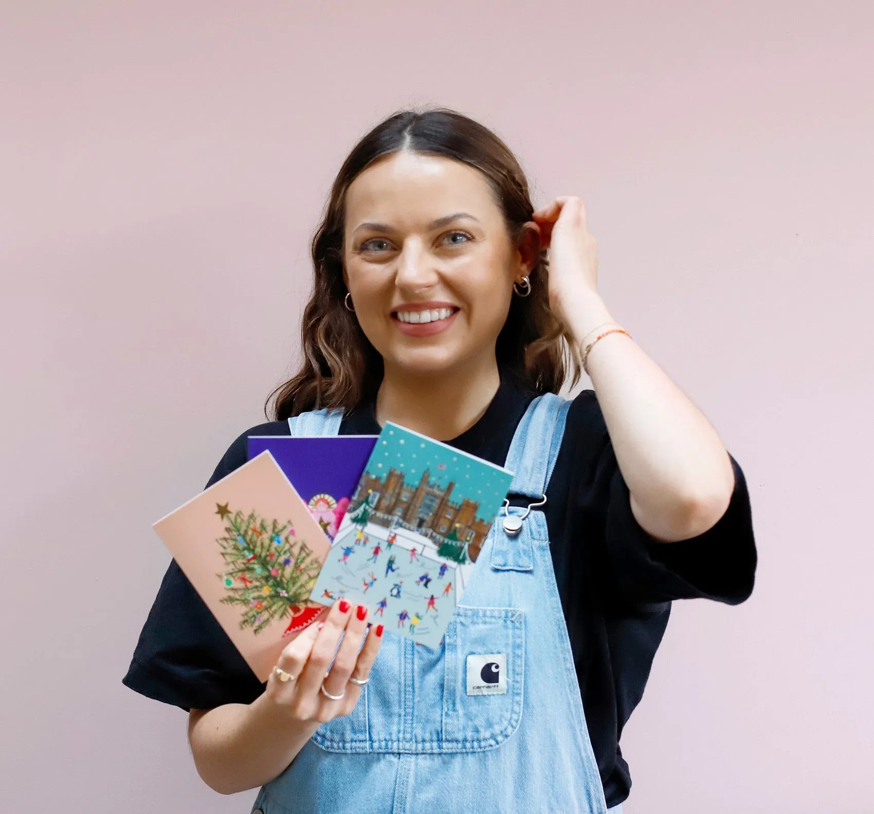 Nush smiling against a pink wall, holding a fan of her illustrated Christmas cards