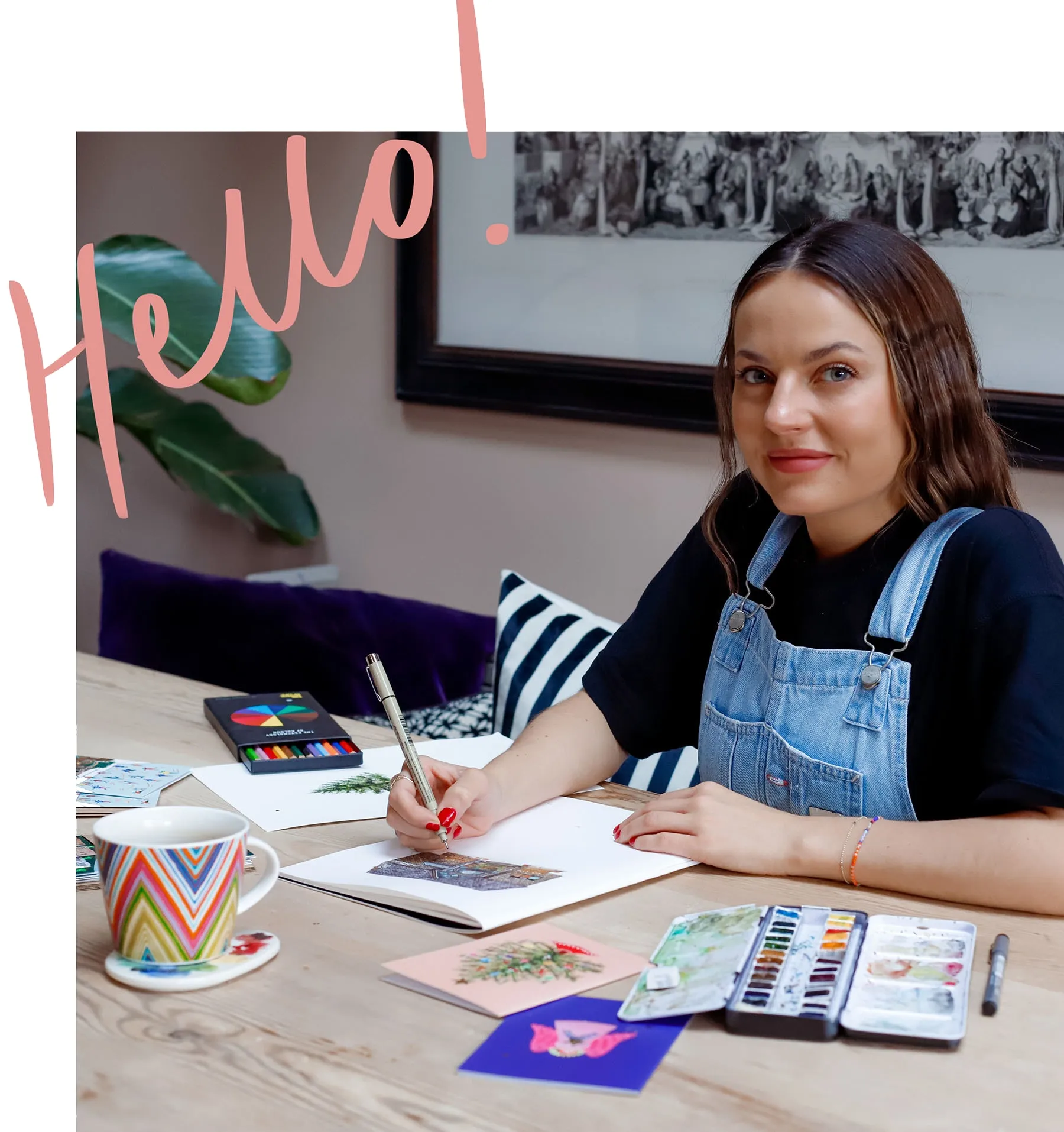 Nush smiling at her desk, drawing with a fineliner, with the word Hello! written beside her