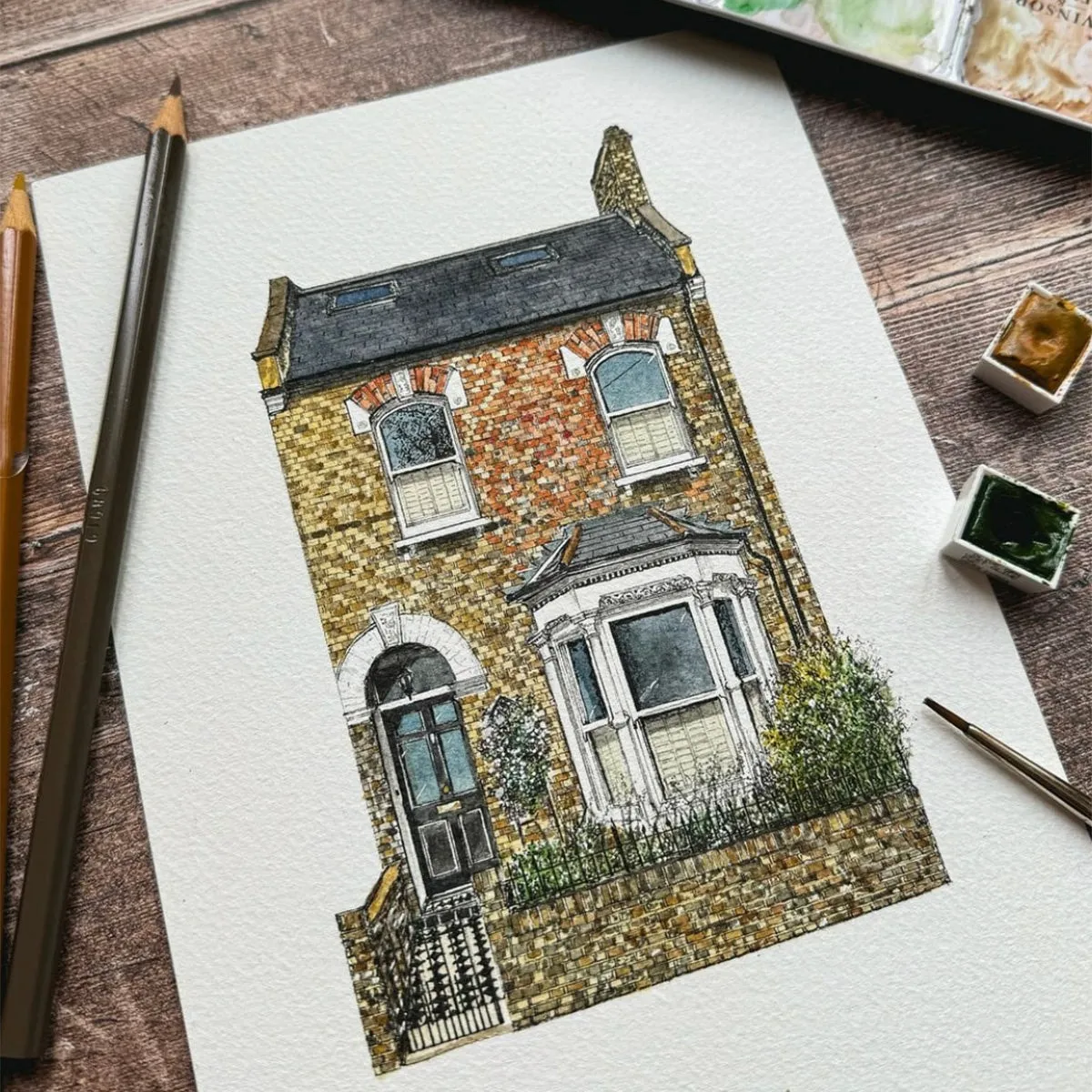 Ink and watercolour house portrait of a red brick Victorian terrace with a bay window, on a wooden table with paints and pencils