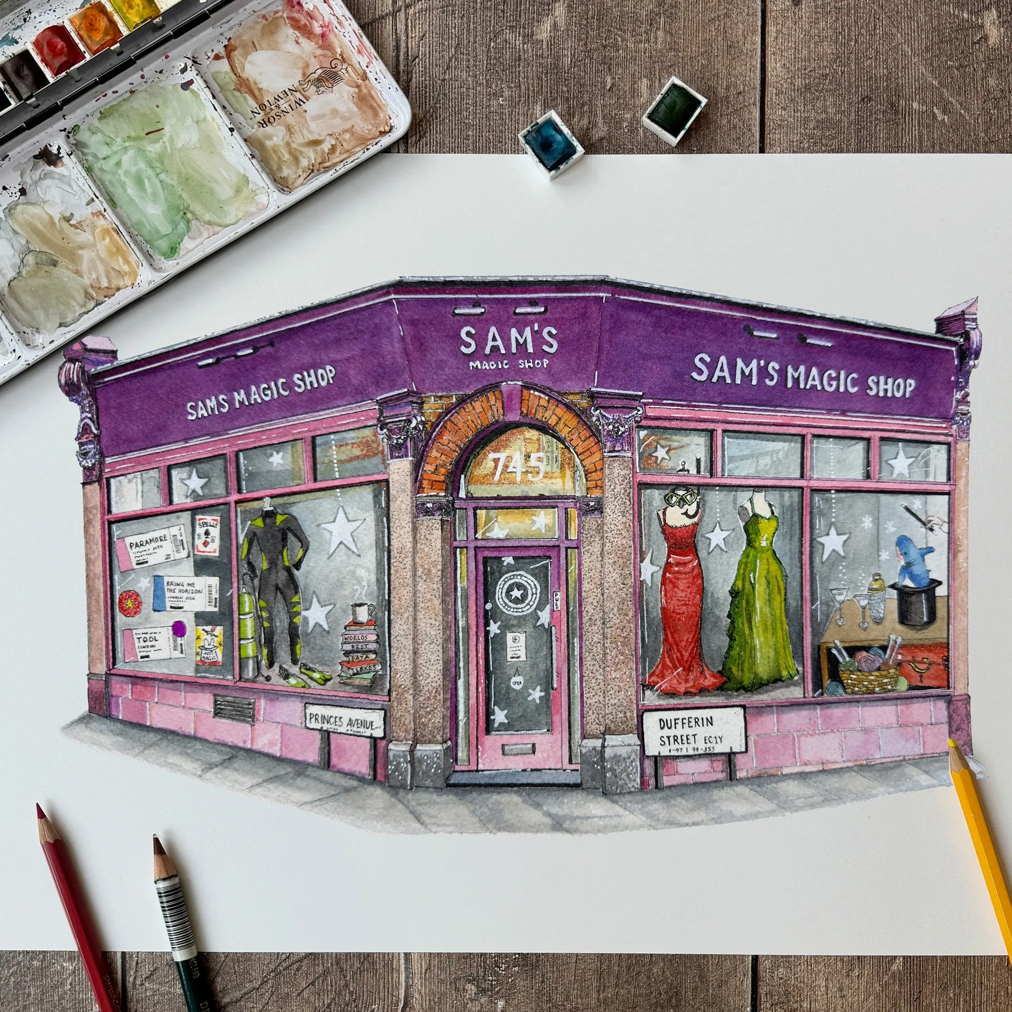 Ink and watercolour drawing of a purple magic shop front with costumes in the windows, on a wooden desk beside a paint palette