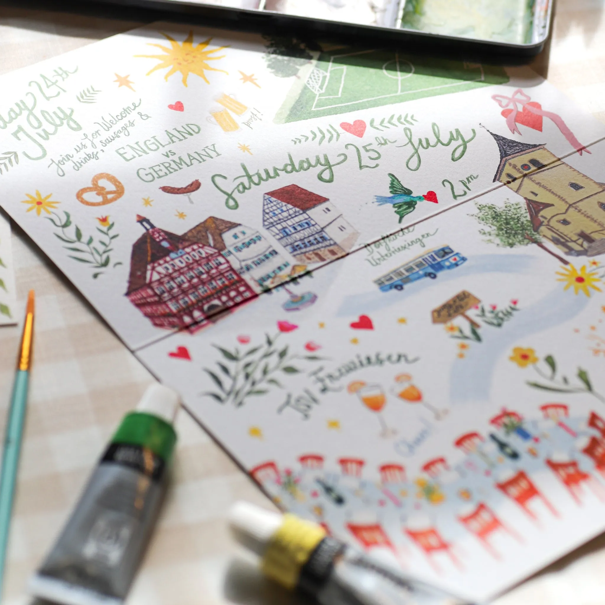Illustrated wedding invitations with colourful hand-drawn buildings, hearts and lettering, surrounded by paint tubes and brushes