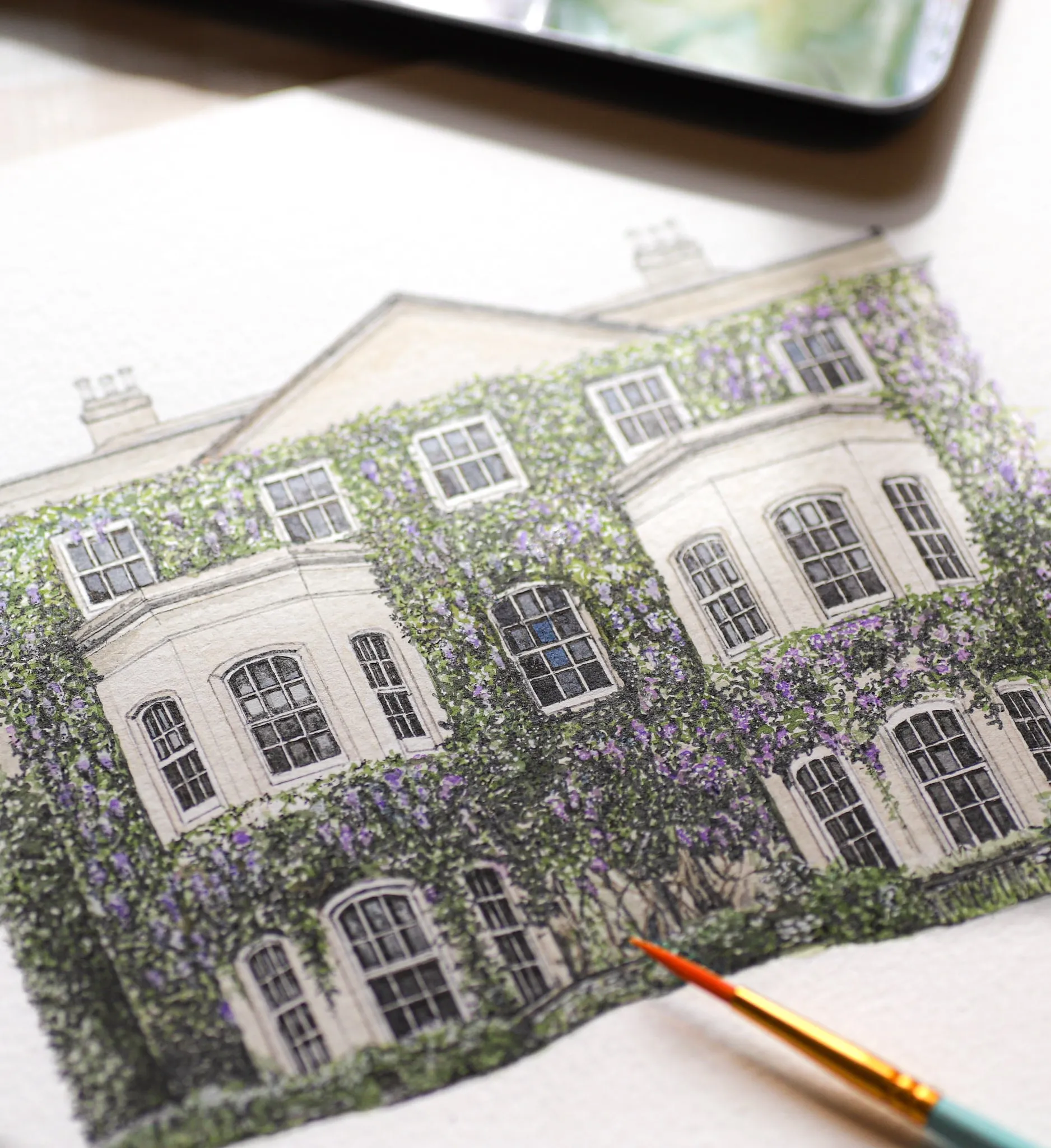 Close-up of an ink and watercolour drawing of a manor house covered in purple wisteria, with a paintbrush resting on the paper
